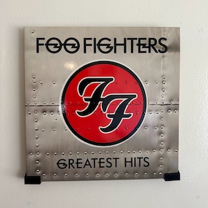 May include: A silver and red album cover with the text "Foo Fighters" and "Greatest Hits". The Foo Fighters logo is a red circle with a black "FF" inside.