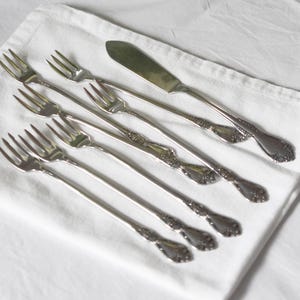 May include: A collection of silver forks and a butter knife arranged on a white napkin. The silverware has ornate detailing on the handles. A clear glass bowl with white flowers sits in the background.