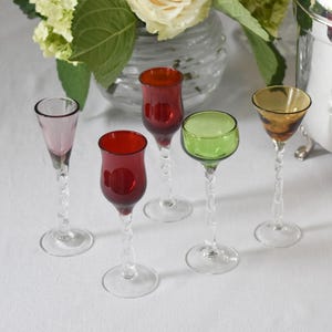 May include: Five colorful glass shot glasses with twisted stems on a white tablecloth. The glasses are red, green, pink, and amber.