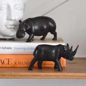 May include: A decorative shelf arrangement featuring a white ceramic face vase with dried foliage, two dark bronze animal figurines, and books. The figurines are a hippopotamus and a rhinoceros. The books have orange and white covers.