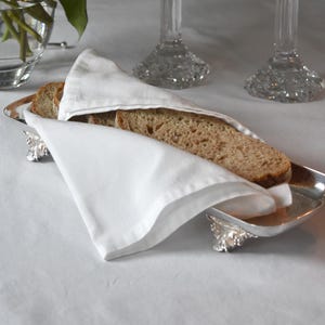 May include: A silver bread basket with a white linen napkin, filled with slices of bread. Two crystal candle holders with white candles are in the background, along with a vase of white roses. The scene is set on a white tablecloth.