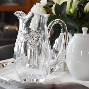 May include: A clear crystal pitcher with a diamond pattern, a curved handle, and a pouring spout. The pitcher sits on a silver tray with a white cloth, alongside a white teapot and a bouquet of white and yellow flowers.