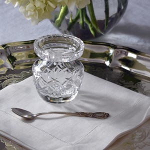 May include: A clear crystal salt cellar with a matching lid sits on a white linen napkin, accompanied by a small silver spoon. The salt cellar has a diamond pattern. A silver tray and a vase of white flowers are in the background.