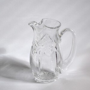 May include: A clear glass pitcher with a decorative diamond pattern and a curved handle. The pitcher has a spout for easy pouring and sits on a white surface. The pitcher is likely used for serving beverages.