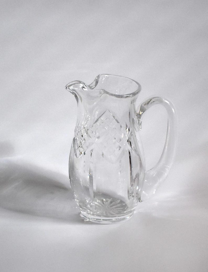 May include: A clear glass pitcher with a decorative diamond pattern and a curved handle. The pitcher has a spout for easy pouring and sits on a white surface. The pitcher is likely used for serving beverages.