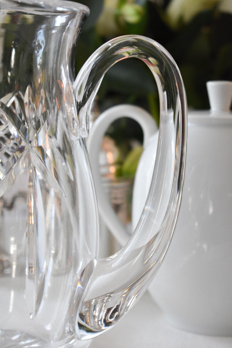 May include: Close-up of a clear glass pitcher with a curved handle. The pitcher has a diamond-cut pattern on the front and a smooth, rounded handle. A white teapot is visible in the background.