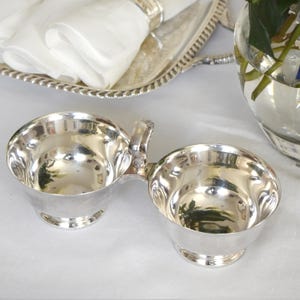 May include: A silver-plated double bowl with a handle, ideal for serving condiments. The bowls have a rounded shape and sit on a small pedestal base. The reflective surface of the bowls creates a bright, elegant appearance. A silver serving tray and a glass vase are in the background.