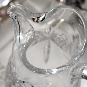 May include: Clear glass pitcher with a curved handle and a decorative starburst pattern on the base. The pitcher has a wide opening and a curved spout. The glass is clear, allowing light to pass through.