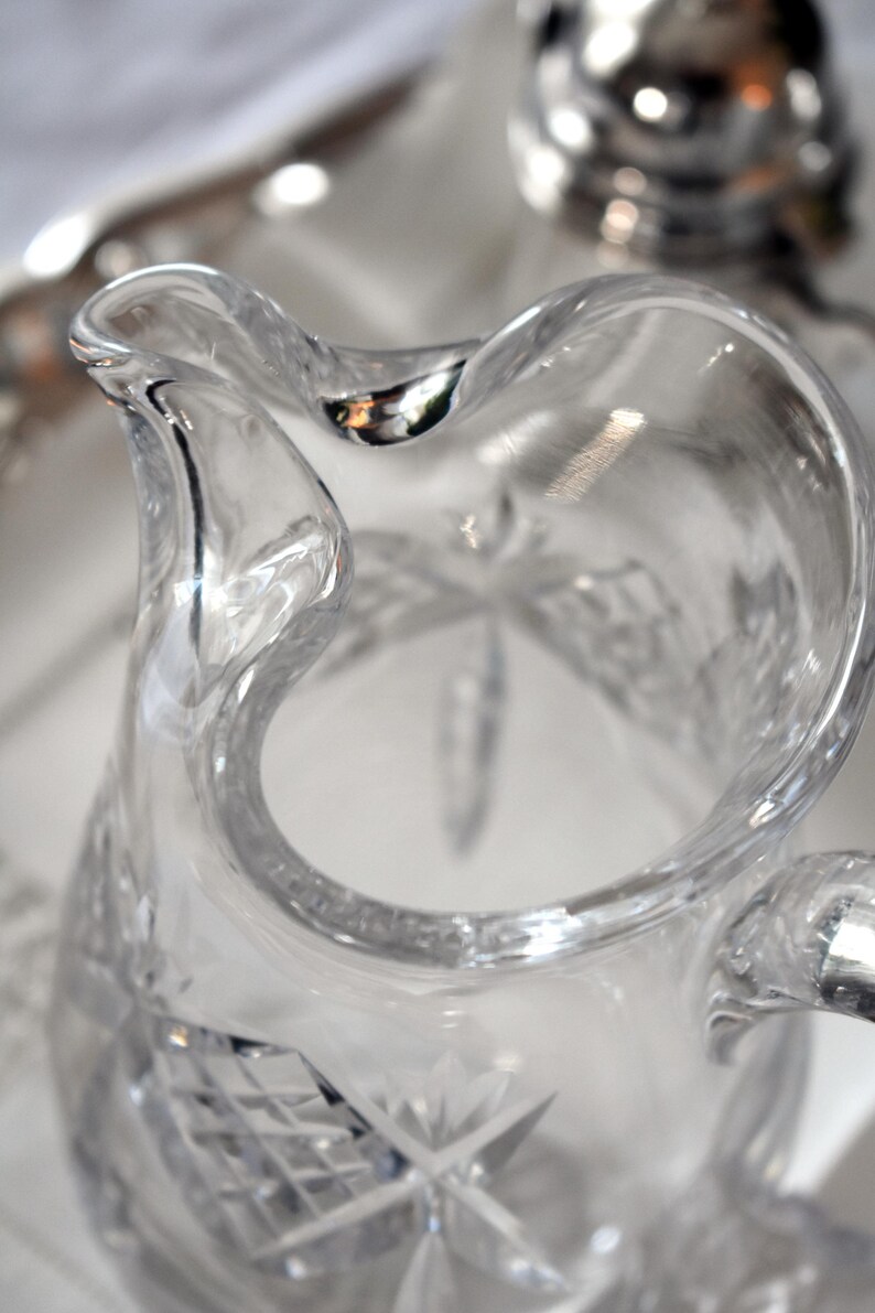 May include: Clear glass pitcher with a curved handle and a decorative starburst pattern on the base. The pitcher has a wide opening and a curved spout. The glass is clear, allowing light to pass through.