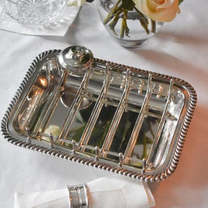 May include: A silver-plated toast rack with a rectangular shape and a decorative rope-like border. The rack has multiple slots for holding toast and a small, round handle. A white napkin with a silver napkin ring is in the foreground.