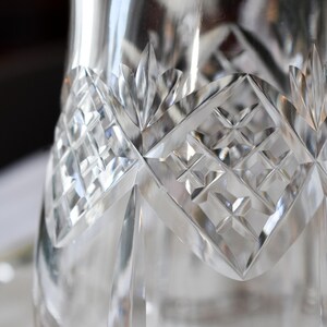 May include: Close-up of a clear, cut-glass pitcher. The pitcher features a diamond pattern and leaf-shaped accents. The glass reflects light, creating a sparkling effect. The design is intricate and detailed.