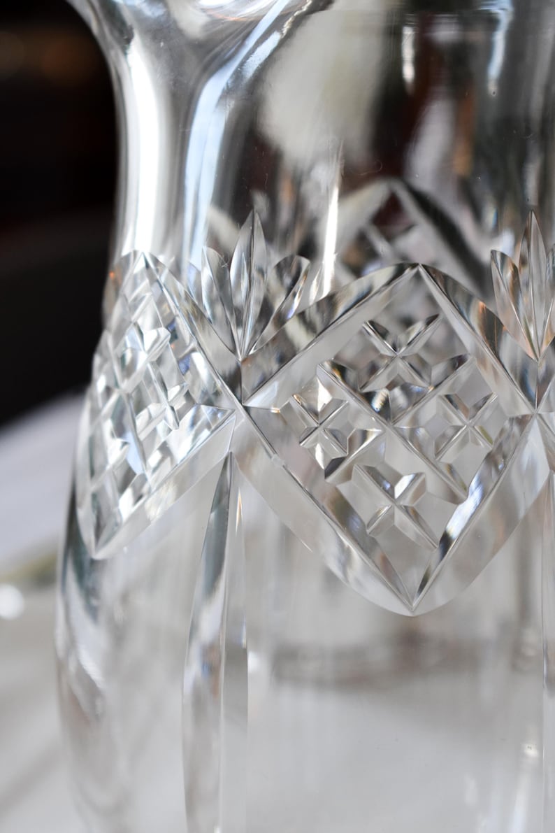 May include: Close-up of a clear, cut-glass pitcher. The pitcher features a diamond pattern and leaf-shaped accents. The glass reflects light, creating a sparkling effect. The design is intricate and detailed.