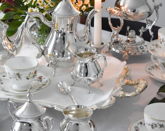 May include: A silver tea set with a teapot, creamer, sugar bowl, and two spoons on a white tablecloth. The tea set is on a silver tray with a candle in the background.