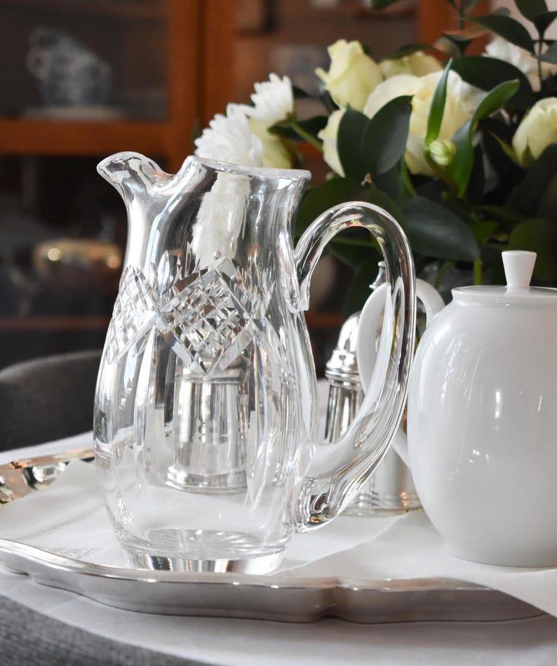 May include: A clear crystal pitcher with a diamond pattern, a curved handle, and a pouring spout. The pitcher sits on a silver tray with a white cloth, alongside a white teapot and a bouquet of white and yellow flowers.