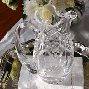 May include: A clear, cut-glass pitcher with a handle, sitting on a silver tray with a white linen napkin. The pitcher has a diamond pattern and is next to a bouquet of white flowers.