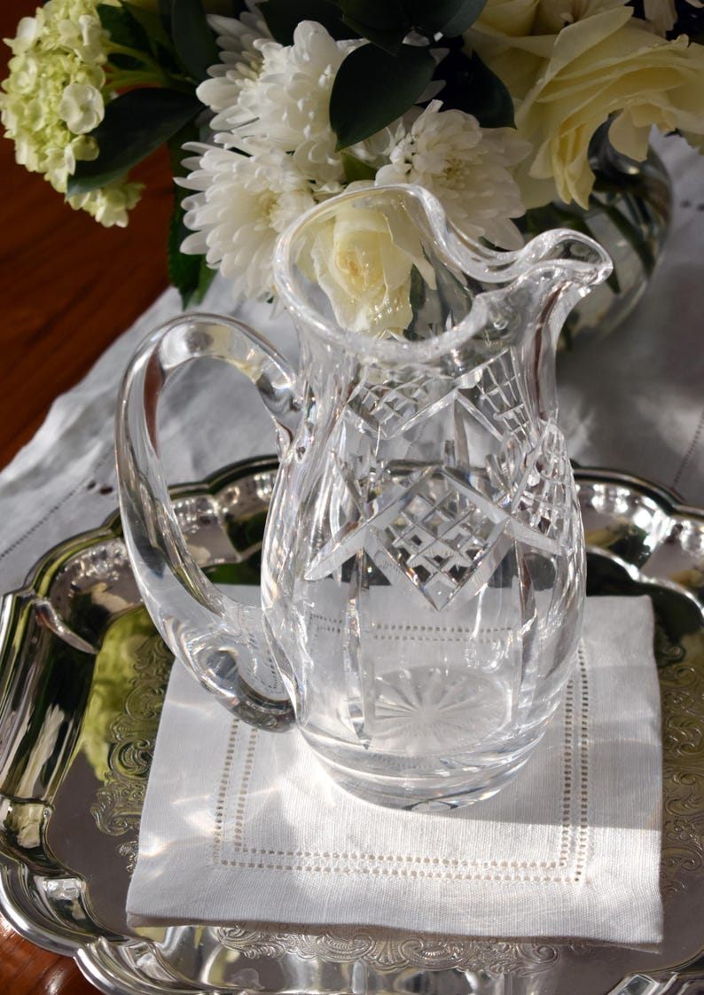 May include: A clear, cut-glass pitcher with a handle, sitting on a silver tray with a white linen napkin. The pitcher has a diamond pattern and is next to a bouquet of white flowers.