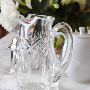 May include: A clear, cut-glass pitcher with a diamond pattern, featuring a curved handle and a pouring spout. The pitcher is part of a table setting, with a white sugar bowl and a bouquet of white flowers in the background.