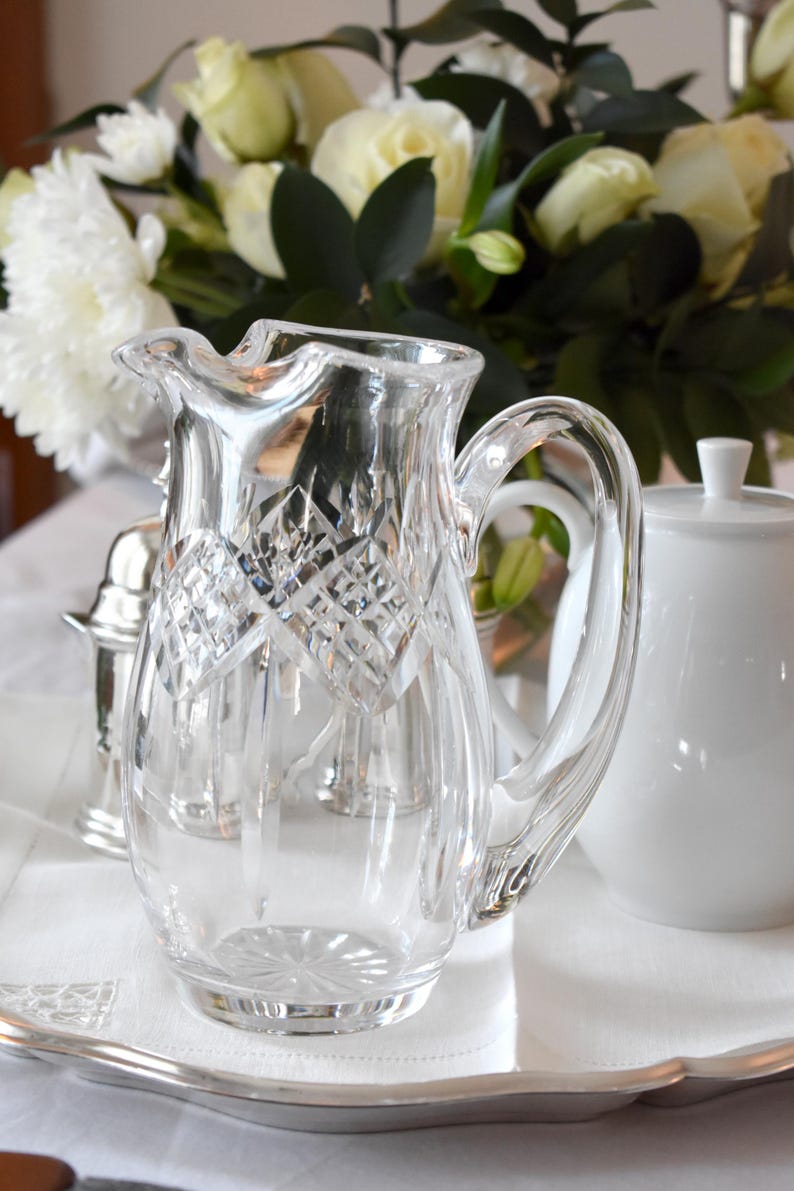 May include: A clear, cut-glass pitcher with a diamond pattern, featuring a curved handle and a pouring spout. The pitcher is part of a table setting, with a white sugar bowl and a bouquet of white flowers in the background.