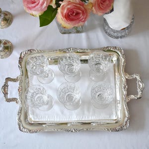 May include: A silver serving tray with six crystal glasses, a bouquet of pink roses, and a bottle of champagne in a crystal ice bucket. The tray has ornate handles and a decorative border.