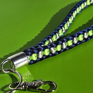May include: A braided wrist strap keychain in navy blue, white, and neon green. The keychain has a silver-colored metal clasp and key ring. The strap is made of a woven cord, and the background is a solid green.