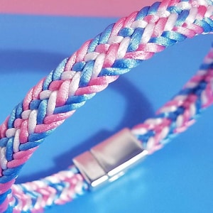 May include: Braided bracelet featuring pink, white, and blue threads. The bracelet has a magnetic clasp and a woven design. The color scheme is a mix of pastel and bright hues, creating a visually appealing accessory.