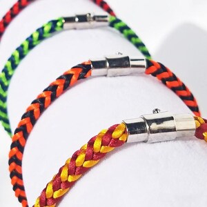 Op de afbeelding: Close-up van verschillende gevlochten armbanden in verschillende kleuren, waaronder rood, geel, groen en oranje. Elke armband heeft een zilveren magnetische sluiting. De armbanden worden op een wit oppervlak getoond.