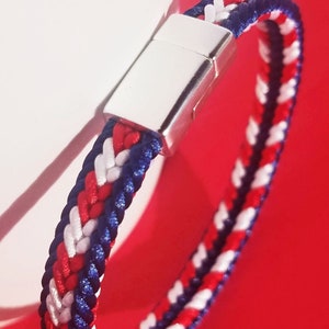 Könnte beinhalten: Ein geflochtenes Armband mit roten, weißen und blauen Fäden. Das Armband hat einen silbernen Magnetverschluss.