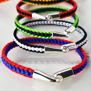 Op de afbeelding: Een stapel kleurrijke gevlochten armbanden met zilveren sluitingen. De armbanden zijn rood, blauw, wit, zwart, groen, geel en oranje.