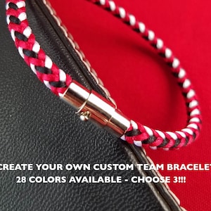 May include: Braided bracelet in red, white, and black with a silver magnetic clasp. The bracelet is on a black and red background. Text at the bottom reads "CREATE YOUR OWN CUSTOM TEAM BRACELET 28 COLORS AVAILABLE - CHOOSE 3!!!"