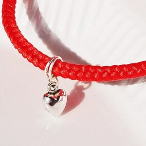 May include: A red string bracelet with a silver heart charm. The bracelet is made of a braided cord and the heart charm is attached to a silver ring.