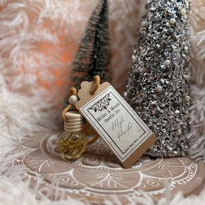 May include: A small, clear glass bottle with a wooden cap and a beige tag that reads "Wicks & Woods Candle Co. Maple Pancakes". The bottle is suspended by a beige string. Two decorative Christmas trees are in the background.