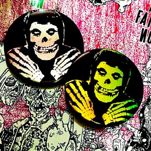 以下が含まれることがあります： 笑顔の頭蓋骨の緑と黄色の画像が描かれた、黒と白の丸いボタンが2つ。頭蓋骨は黒いヘルメットをかぶっており、緑と黄色の背景があります。