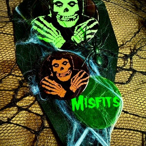 Puede incluir: Tres pines redondos con fondo verde, naranja y negro. Los pines presentan una calavera con un diseño verde, naranja y negro. El pin inferior tiene la palabra "Misfits" escrita en verde.