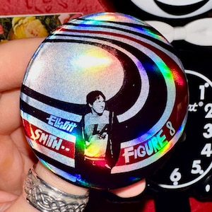 Puede incluir: Un botón plateado y negro con acabado holográfico. El botón presenta una imagen en blanco y negro de una persona con el texto "Elliot Smith - Figure 8" en blanco.