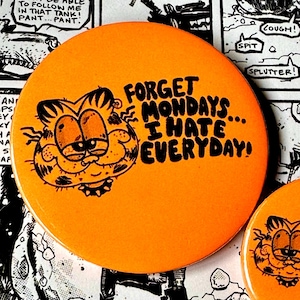 Może przedstawiać: Dwa pomarańczowe i czarne guziki Garfield z napisem "Forget Mondays... I Hate Everyday!" na nich.