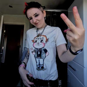May include: White t-shirt featuring a cartoon illustration of a baby with spiky orange hair, a black vest, and jeans. The baby is giving a peace sign. The person is wearing multiple necklaces and a watch.