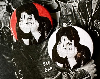 May include: Two black and white button pins with a red background on one. The pins feature a woman with her middle fingers up in front of her face. The text "SID RIP" is written on the bottom of one pin.