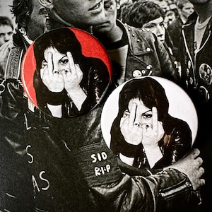 Può includere: Due spille da balia in bianco e nero con uno sfondo rosso su una. Le spille raffigurano una donna con le dita medie alzate davanti al viso. Il testo "SID RIP" è scritto sul fondo di una spilla.