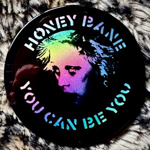 Może przedstawiać: Czarno-biały guzik z holograficznym obrazem twarzy osoby w tęczowych kolorach. Wokół obrazu nadrukowany jest tekst "Honey is Rave" i "You Can Be You".