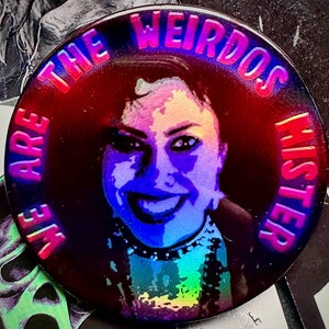 Puede incluir: Un botón redondo con fondo negro y una imagen arcoíris de la cara de una mujer. El texto "We Are The Weirdos Mister" está escrito en rosa alrededor del borde del botón.