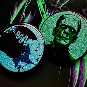 May include: Two round pins with a glittery blue and green finish. One pin features a black and white silhouette of Frankenstein's monster. The other pin features a black and white silhouette of a woman with a hairstyle reminiscent of Frankenstein's monster.