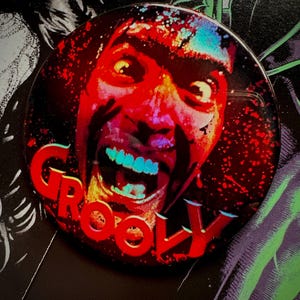 Könnte beinhalten: Ein runder Button mit einem rot-schwarzen Design, das ein schreiendes Gesicht mit blauen Zähnen und das Wort "GROOVY" in roten Buchstaben zeigt. Der Button hat eine glänzende Oberfläche und ist vor einem dunklen Hintergrund platziert.