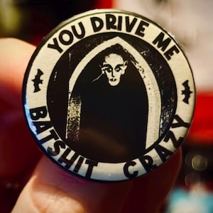 May include: A round, black and white button with the text "YOU DRIVE ME BATSHIT CRAZY" encircling a central image. The image features a stylized figure in a dark archway. Small bat silhouettes are on the sides.