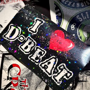May include: A black sticker with the text "I D-BEAT" in white, outlined in black, and a red heart. The sticker has a holographic, glittery effect with colorful stars and specks. Other stickers are visible in the background.