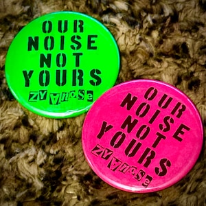 Może przedstawiać: Dwa okrągłe guziki z tekstem "OUR NOISE NOT YOURS" czarnymi literami na zielonym i różowym tle. Tekst "ZYnoise" jest również widoczny na obu guzikach.
