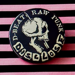 Pode incluir: Botão de metal preto e branco com um design de caveira e ossos cruzados. O texto no botão diz "D-Beat Raw Punk Disclose".
