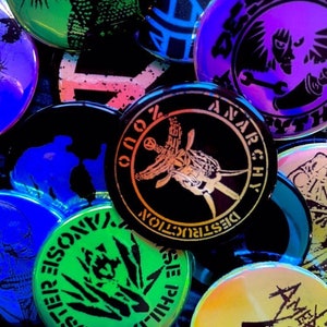 Puede incluir: Una colección de pines redondos de colores con varios diseños gráficos. Los pines presentan ilustraciones en blanco y negro de animales, calaveras y otros diseños abstractos. Algunos de los pines tienen texto, incluyendo "Destruction", "Anarchy" y "Crisis".
