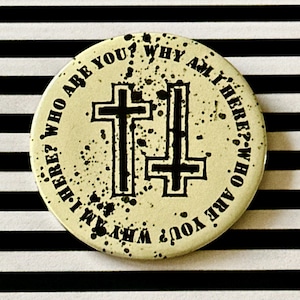 May include: A white button with black text and a black graphic of two crosses. The text reads "Who are you? Why am I here? Who are you? Why am I here?"