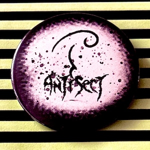 Könnte beinhalten: Ein schwarz-weißer Knopf mit rosa Hintergrund. Der Knopf zeigt eine stilisierte Grafik eines Schädels mit dem Wort "Antisect" darunter.