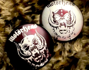 May include: Two round, black and white, pinback buttons with the band name "Motorhead" and a snarling, horned, animal head graphic.